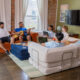 Living room with couches around an ottoman with 5 young men hanging out.