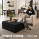 Woman in white chair with feet up resting on black foam ottoman with cat on back legs resting fronting legs on ottoman.