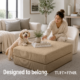 Woman in white chair picking up cup off of cream ottoman with dog on back legs resting fronting legs on ottoman.
